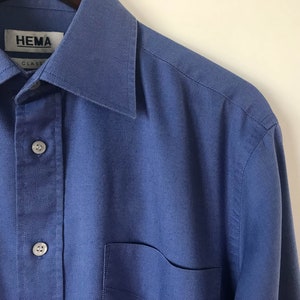 May include: A blue long-sleeved button-down shirt with a pocket on the left side. The shirt has a label that reads "HEMA CLASS".