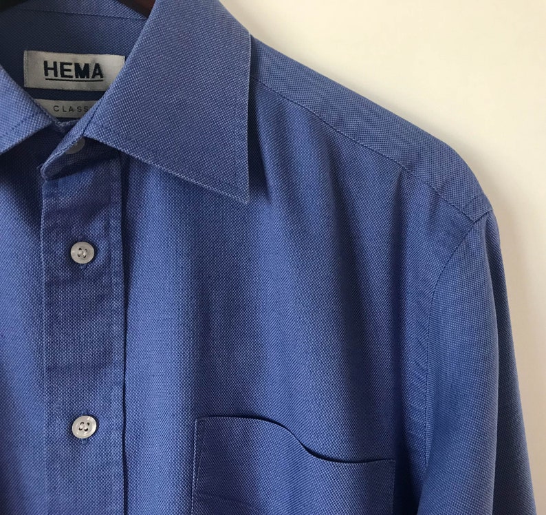 May include: A blue long-sleeved button-down shirt with a pocket on the left side. The shirt has a label that reads "HEMA CLASS".