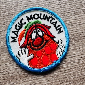 May include: Round embroidered patch with the words "MAGIC MOUNTAIN" in black around the edge. The patch features a cartoon character with a red body, green hair, and white hands. The patch is bordered with blue stitching.