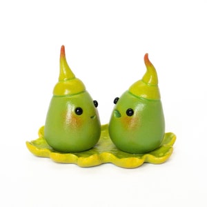 Peut inclure: Deux figurines en céramique vertes avec des chapeaux jaunes et des yeux noirs, assises sur une base en forme de feuille verte.