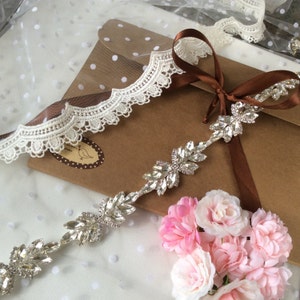May include: A delicate rhinestone trim with a leaf design, featuring clear crystals. The trim is displayed on a brown paper background with white polka dots, along with a brown satin ribbon and a white lace trim.