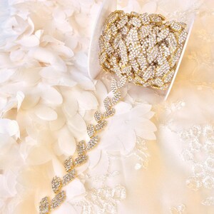 May include: Gold rhinestone trim with a leaf design. The trim is on a white fabric background.