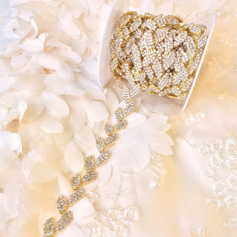 May include: Gold rhinestone trim with a leaf design. The trim is on a white fabric background.