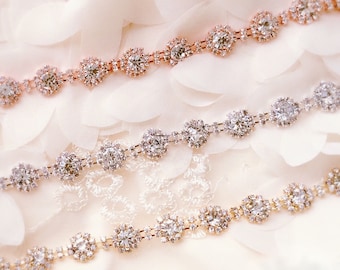 May include: Three rows of rhinestone trim with gold and rose gold settings. The trim is on a white fabric background.