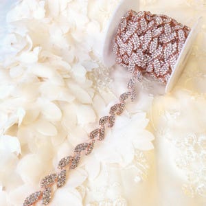 May include: A spool of rose gold rhinestone trim with a leaf design. The trim is on a white fabric background.