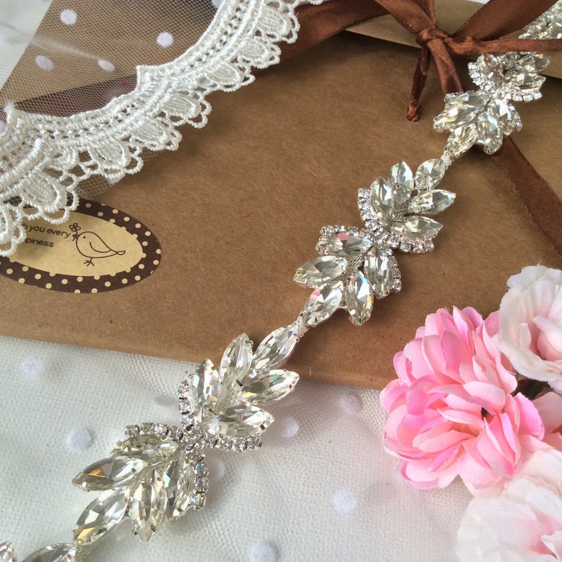 May include: A silver rhinestone trim with large, clear, faceted rhinestones arranged in a leaf pattern. The trim is on a brown background with white lace and pink flowers.