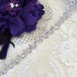 May include: A close-up of a bridal headpiece featuring a deep purple fabric flower with a black veil and a sparkling rhinestone trim. The trim is made of clear, oval-shaped rhinestones set in a silver metal.