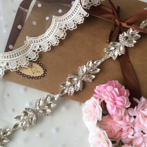 May include: A silver rhinestone trim with a leaf design. The trim is on a brown background with white lace and pink flowers.