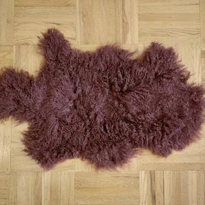 May include: A plush, mauve-colored sheepskin rug with long, curly fur. The rug is laid out flat on a wooden floor.