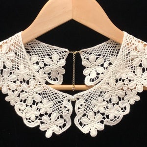 May include: White lace collar with a delicate floral pattern. The collar has a chain attached to the back for fastening.