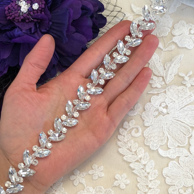 May include: A silver-toned, leaf-shaped rhinestone trim. The trim features clear, faceted rhinestones set in a delicate, repeating pattern. The design is held in a hand, with a purple flower and lace in the background.