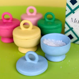 May include: A selection of colourful ceramic salt cellars and lids in pink, yellow, green, and blue. One blue cellar contains white salt. Each cellar has a matching lid with a ring handle. The background is a bright green.