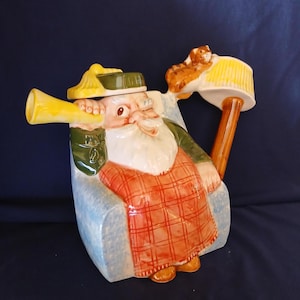 May include: A ceramic teapot shaped like a seated man with a white beard, wearing a green hat and jacket, and a red plaid kilt. The teapot has a yellow horn and a squirrel on the lid. The teapot is on a blue background.