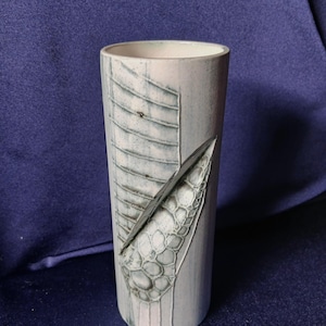 May include: A cylindrical ceramic vase with a light gray and white color scheme. The vase features textured, geometric designs on the exterior, including a series of parallel lines and a scale-like pattern. The vase has a smooth, rounded rim.