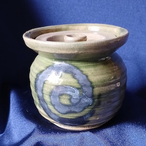Puede incluir: Un tarro de cerámica hecho a mano con tapa. El tarro tiene forma redondeada con una abertura ancha y una tapa con un agujero central. El exterior está esmaltado en tonos verdes y beige, con un diseño en espiral azul y gris. El tarro mide aproximadamente 10 cm de alto.