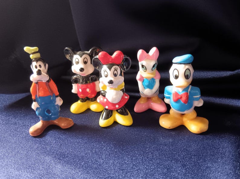 May include: A collection of five Disney character figurines. Goofy in orange and blue, Mickey Mouse in red and black, Minnie Mouse in a red polka dot dress, Daisy Duck in pink and blue, and Donald Duck in blue. The figurines are on a dark blue surface.