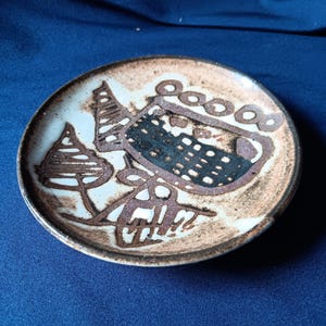 May include: A small, round ceramic plate with a brown and white abstract design. The plate features a dark brown rim and a central design with geometric shapes and lines. The plate is set against a blue background.