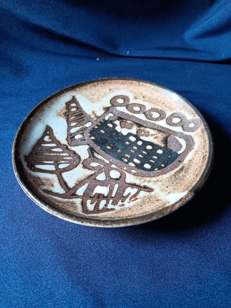May include: A small, round ceramic plate with a brown and white abstract design. The plate features a dark brown rim and a central design with geometric shapes and lines. The plate is set against a blue background.
