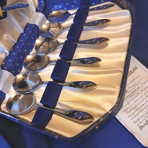 May include: A collection of silver spoons with starburst patterns on the handles, presented in a blue velvet-lined case. The spoons rest on cream satin, secured by blue velvet straps. The case is blue with a white star design. The case is blue.