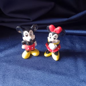 May include: Two ceramic figurines of Mickey and Minnie Mouse. Mickey is wearing red shorts with white polka dots, yellow shoes, and a black body. Minnie is wearing a red polka dot skirt, yellow shoes, and a red bow.