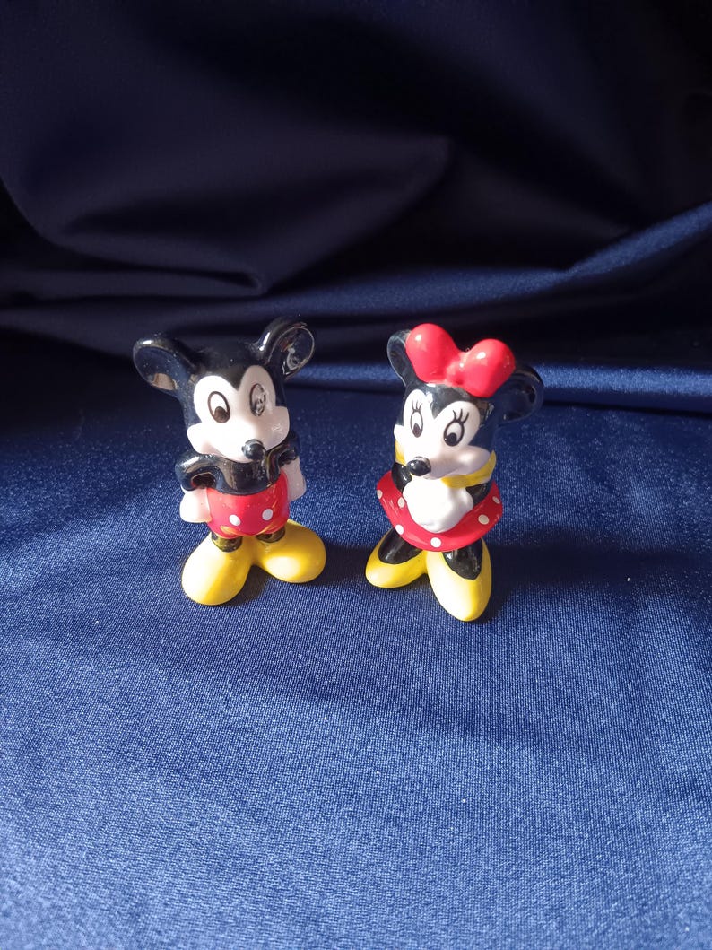 May include: Two ceramic figurines of Mickey and Minnie Mouse. Mickey is wearing red shorts with white polka dots, yellow shoes, and a black body. Minnie is wearing a red polka dot skirt, yellow shoes, and a red bow.