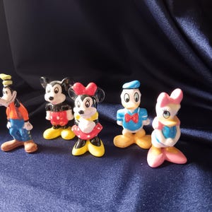 May include: A collection of five Disney character figurines. Goofy in an orange shirt and blue pants, Mickey Mouse in red shorts, Minnie Mouse in a red dress, Donald Duck in a blue shirt, and Daisy Duck in a pink bow.