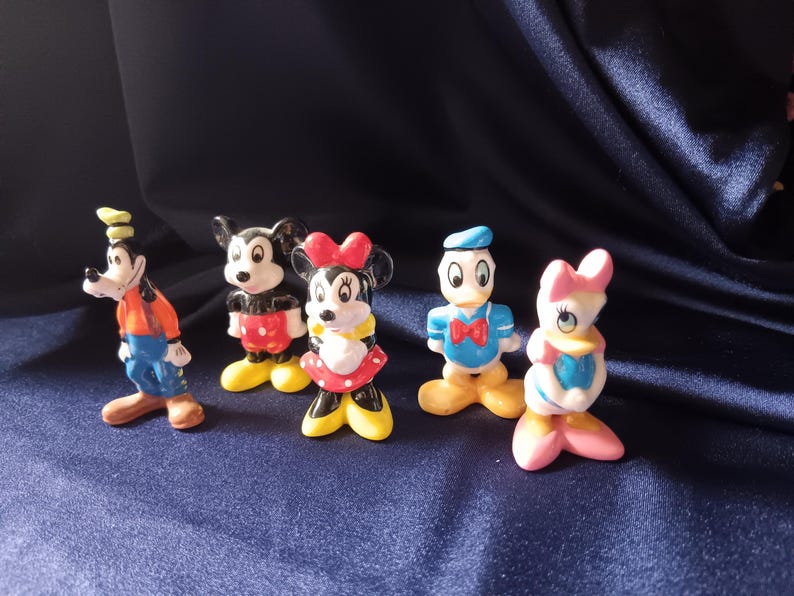 May include: A collection of five Disney character figurines. Goofy in an orange shirt and blue pants, Mickey Mouse in red shorts, Minnie Mouse in a red dress, Donald Duck in a blue shirt, and Daisy Duck in a pink bow.