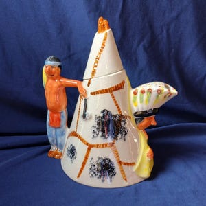 May include: A ceramic teapot in the shape of a Native American teepee, featuring a Native American figure on each side. The teapot is white with brown and black accents, and the figures are painted in bright colors. The spout is shaped like a headdress.