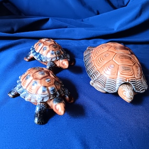 May include: Three ceramic turtle figurines. The largest turtle is brown with a dark brown shell. The two smaller turtles are brown with a blue and brown shell.
