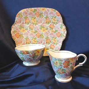 以下が含まれることがあります： 花柄のティーセット。サービングプレート、ボウル、クリーマーが含まれています。セットは、オレンジ、黄色、ピンクの花が咲く水色の背景です。各ピースの縁は金色のトリムで仕上げられています。