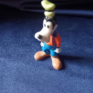 May include: A small figurine of Goofy, a Disney cartoon character. Goofy is wearing a green hat, an orange shirt, blue overalls, and brown shoes. The figurine is standing on a blue surface.