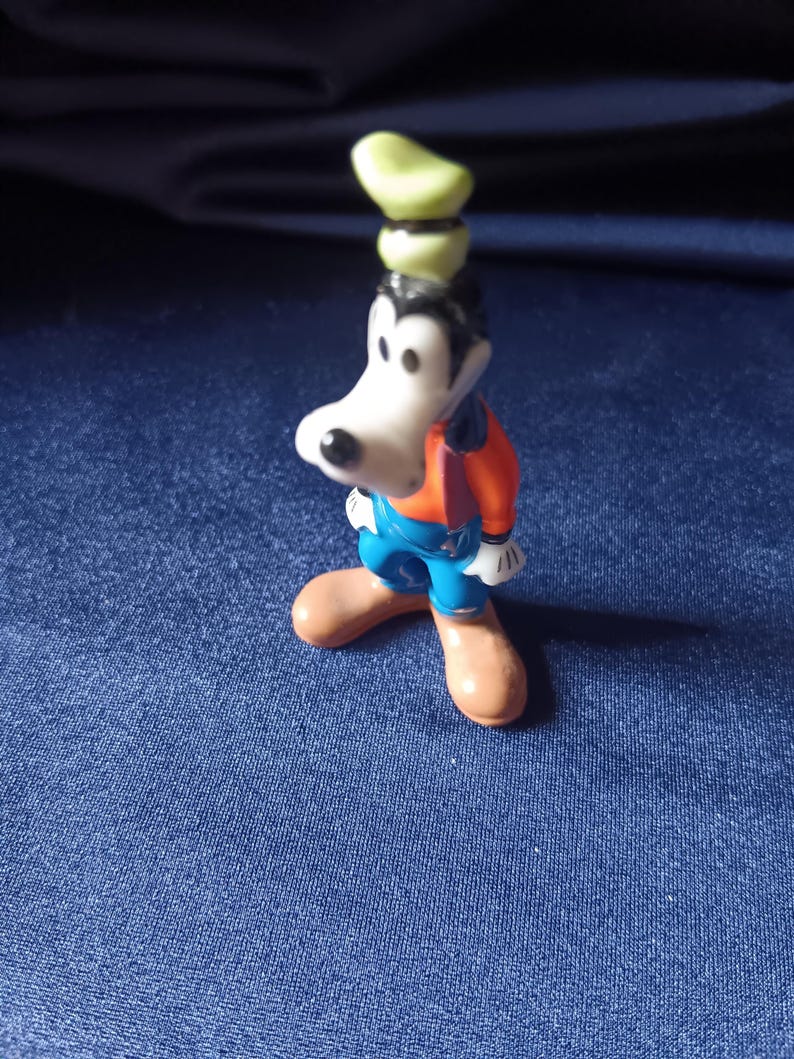 May include: A small figurine of Goofy, a Disney cartoon character. Goofy is wearing a green hat, an orange shirt, blue overalls, and brown shoes. The figurine is standing on a blue surface.