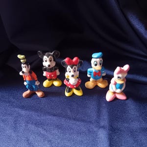 May include: A collection of five Disney character figurines. Mickey Mouse, Minnie Mouse, Donald Duck, Daisy Duck, and Goofy are depicted in their classic outfits. The figurines are brightly colored and stand on a dark blue surface.