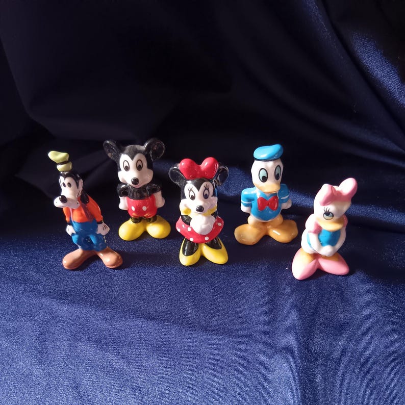 May include: A collection of five Disney character figurines. Mickey Mouse, Minnie Mouse, Donald Duck, Daisy Duck, and Goofy are depicted in their classic outfits. The figurines are brightly colored and stand on a dark blue surface.