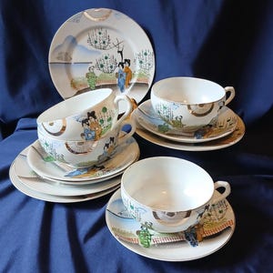 May include: A vintage porcelain tea set featuring a plate and cups with saucers. The set is decorated with a Japanese landscape scene, including figures, trees, and a bridge. The rims of the plates and cups have a gold trim. The background is a deep blue.