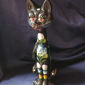 Puede incluir: Figura de gato de cerámica negra con ojos rojos y detalles blancos. El gato está decorado con motivos florales en azul, blanco y verde. La figura tiene un cuello largo y está sentada.