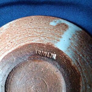 May include: A handmade ceramic bowl with a light blue rim and a brown, textured exterior. The word "ORIGIN" is stamped on the bottom. The bowl has a rustic, artisanal appearance.