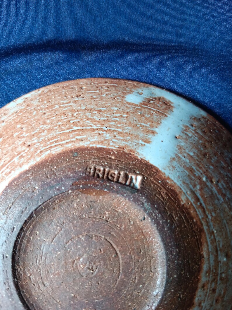 May include: A handmade ceramic bowl with a light blue rim and a brown, textured exterior. The word "ORIGIN" is stamped on the bottom. The bowl has a rustic, artisanal appearance.