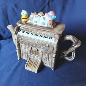 Könnte beinhalten: Eine Keramik-Teekanne in Form eines Klaviers mit brauner Holzmaserung. Auf der Oberseite befinden sich ein offenes Miniatur-Musikbuch, ein kleiner gelber Blumentopf und ein rosa Hut. Sie hat eine weiße Tastatur, goldene Akzente und einen Griff.