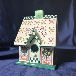 May include: A ceramic house-shaped container with a green base and roof trim. The roof is decorated with floral patterns and the text "Topiary Pot" on a banner. The windows feature potted trees, and the chimney is a small green square.