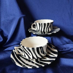 May include: Two ceramic teacups and saucers with a black and white zebra stripe pattern. The cups have square handles and sit on matching saucers. The background is a solid, dark blue fabric.