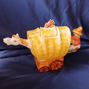 May include: A ceramic teapot shaped like a covered wagon, with a horse head spout and a figure of a person holding the handle. The wagon is yellow and brown, with a small lid and wheels. The teapot has a whimsical, decorative design.