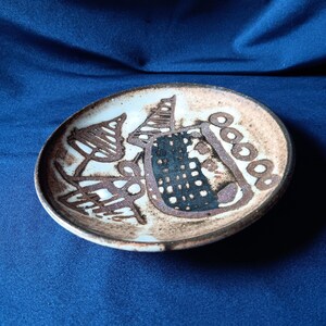 May include: A small, round ceramic plate with a hand-painted design. The plate features a light brown background with abstract brown and black shapes. The rim is a darker brown, and the plate sits on a blue surface.
