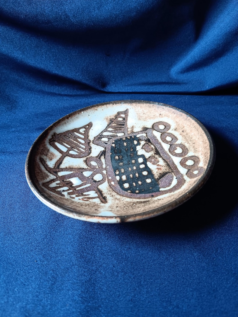 May include: A small, round ceramic plate with a hand-painted design. The plate features a light brown background with abstract brown and black shapes. The rim is a darker brown, and the plate sits on a blue surface.
