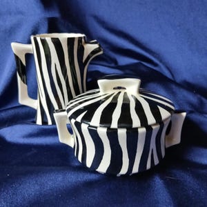 May include: A ceramic creamer and sugar bowl set with a bold black and white zebra stripe pattern. The creamer has a handle and spout, while the sugar bowl has a lid and two small handles. The set is on a blue background.