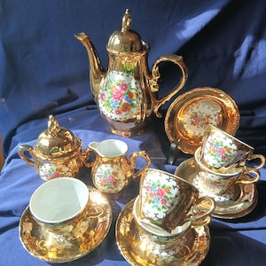 May include: A gold-colored tea set with floral designs. The set includes a teapot, sugar bowl, creamer, cups, and saucers. The floral pattern features pink, blue, and yellow flowers on a white background. The set is arranged on a blue surface.