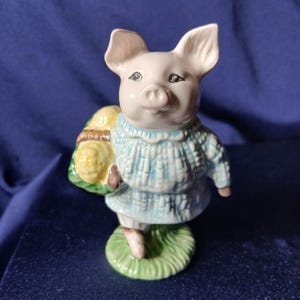 May include: A ceramic pig figurine standing on a green base. The pig is wearing a light blue textured sweater and carrying a small brown basket filled with yellow items. The figurine features painted facial details including eyes and a snout.