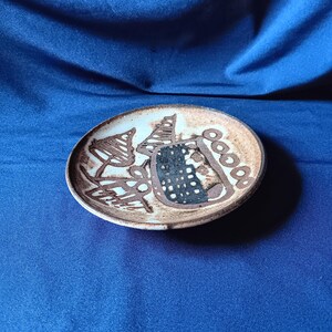 May include: A small, round ceramic plate with a brown rim and a light-colored interior. The plate features abstract brown and black designs, including shapes resembling trees and geometric patterns. The plate is set against a blue background.