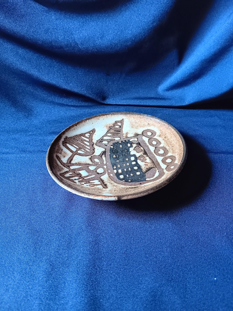 May include: A small, round ceramic plate with a brown rim and a light-colored interior. The plate features abstract brown and black designs, including shapes resembling trees and geometric patterns. The plate is set against a blue background.