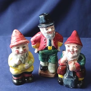 May include: Three ceramic gnome ornaments. One wears a red hat and yellow coat, another a black top hat and red jacket, and the third a red hat and overalls. All have white beards and smiling faces. The ornaments are on a blue background.